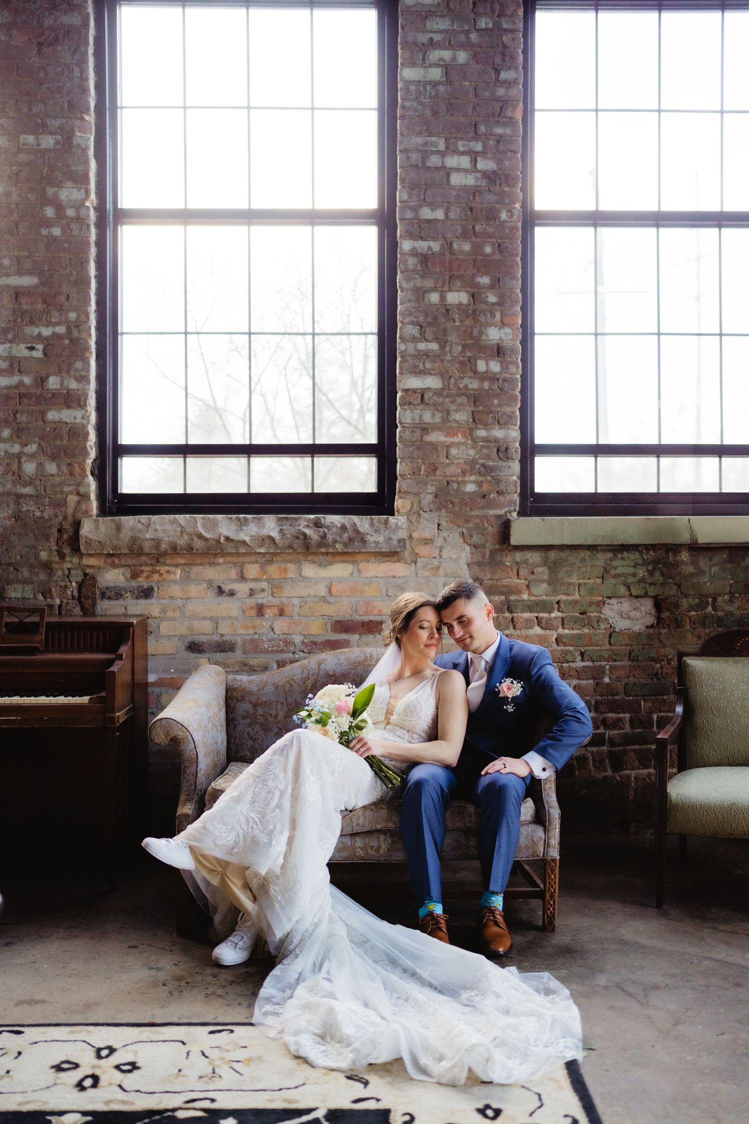 Bride and groom sit together on a vintage sofa in a rustic brick-walled venue with large industrial windows.