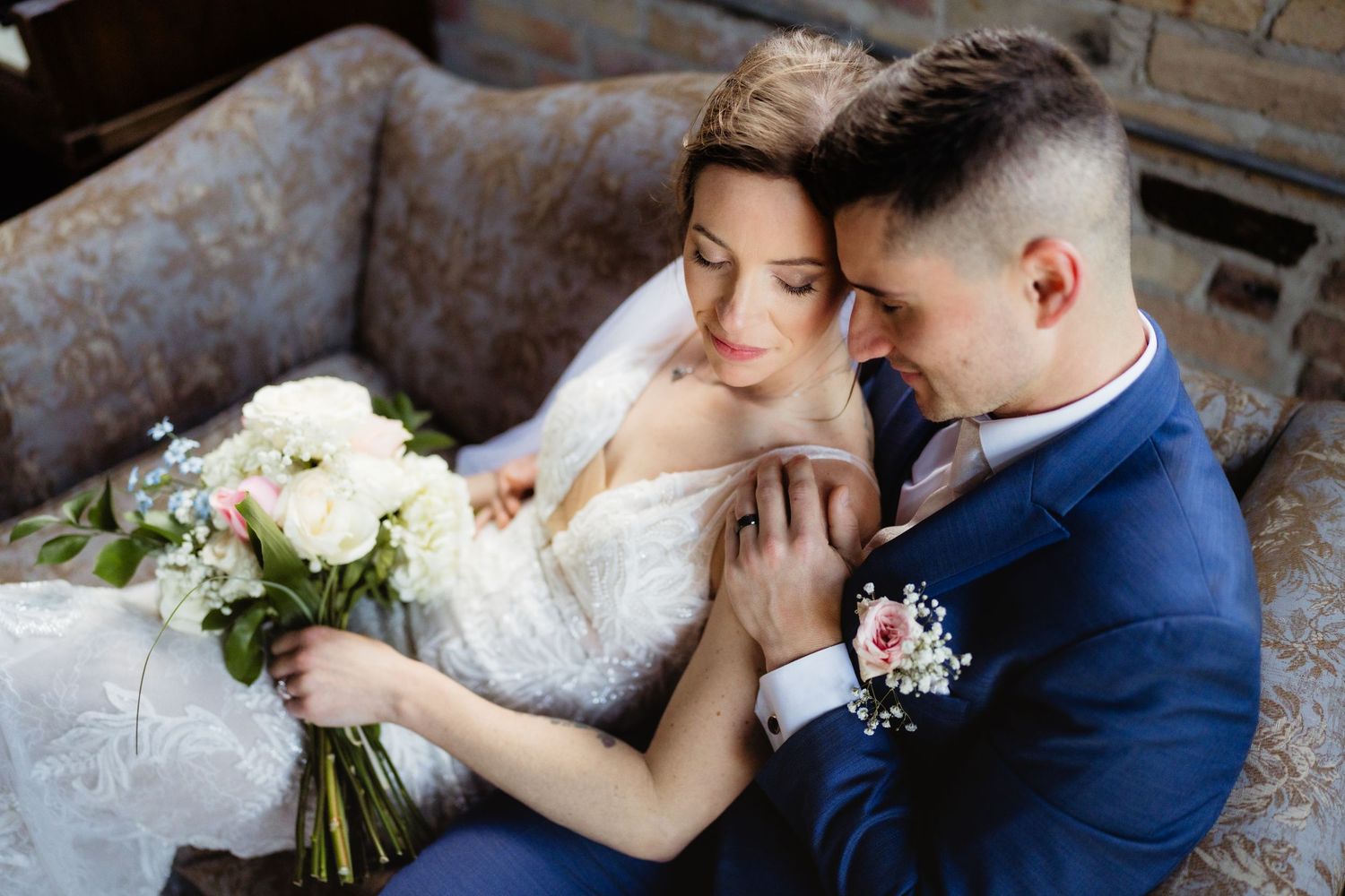 Bride and groom in wedding attire sit closely together, bride holding white floral bouquet, groom in navy suit.