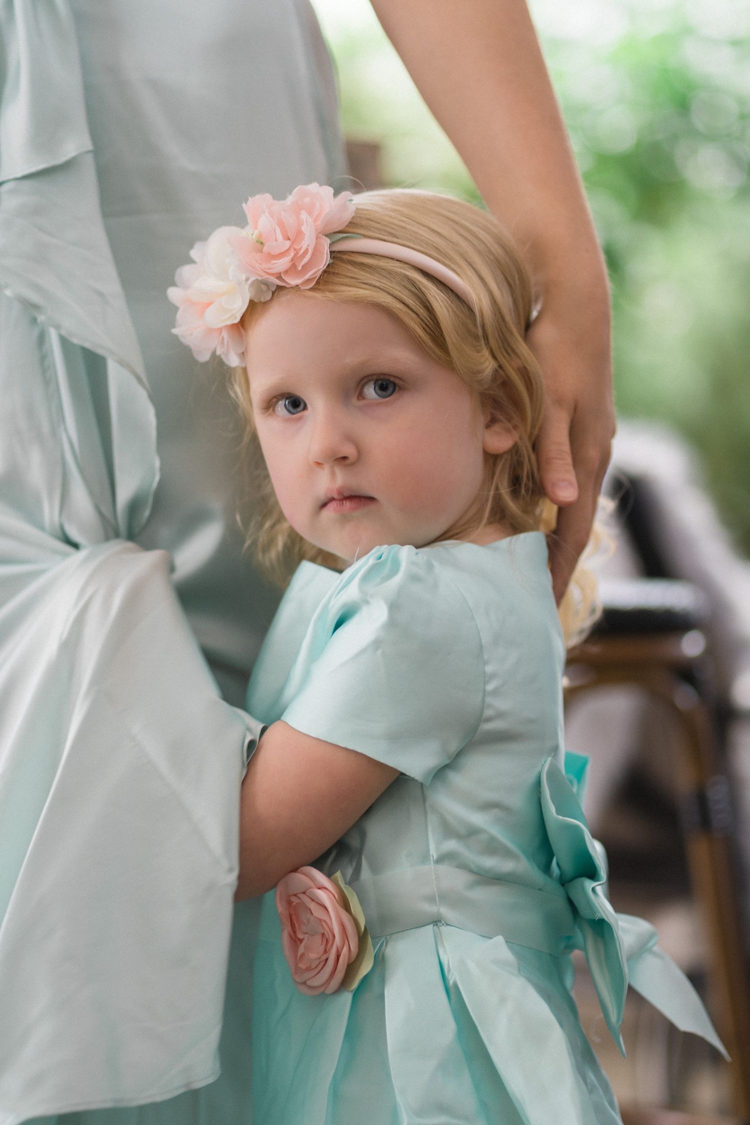 A young child in a mint green dress with peach floral accents stands beside someone in a light-colored dress at an event.