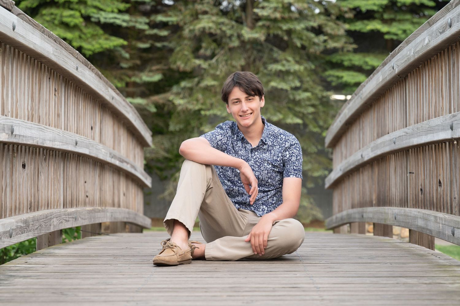 Person sitting casually on a wooden bridge walkway surrounded by lush greenery in a park setting.