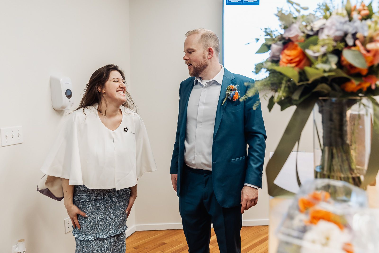 A laughing bride and groom in blue suit share a joyful moment indoors near a floral arrangement.