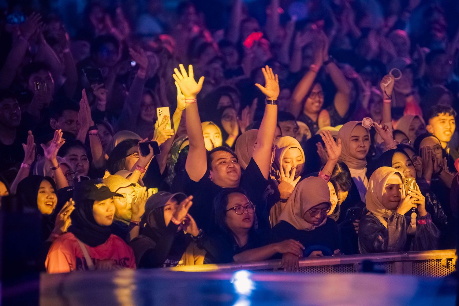 Excited concert crowd with raised hands enjoying a live music event under colorful stage lights.