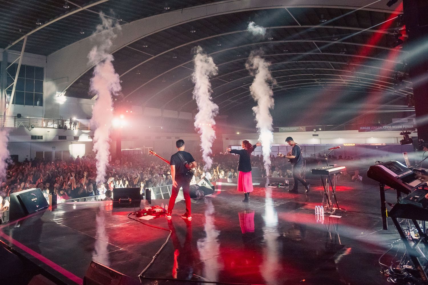 Live concert performance on stage with smoke effects, red lighting, and a large crowd in an indoor venue.