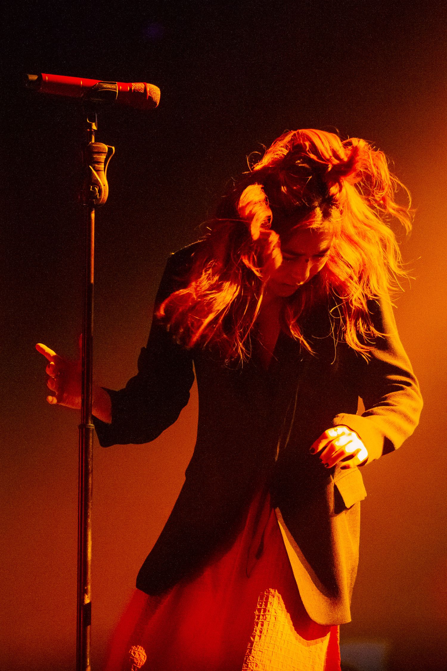 Female singer performing on stage under dramatic orange lighting, holding a microphone stand while her hair flows freely.