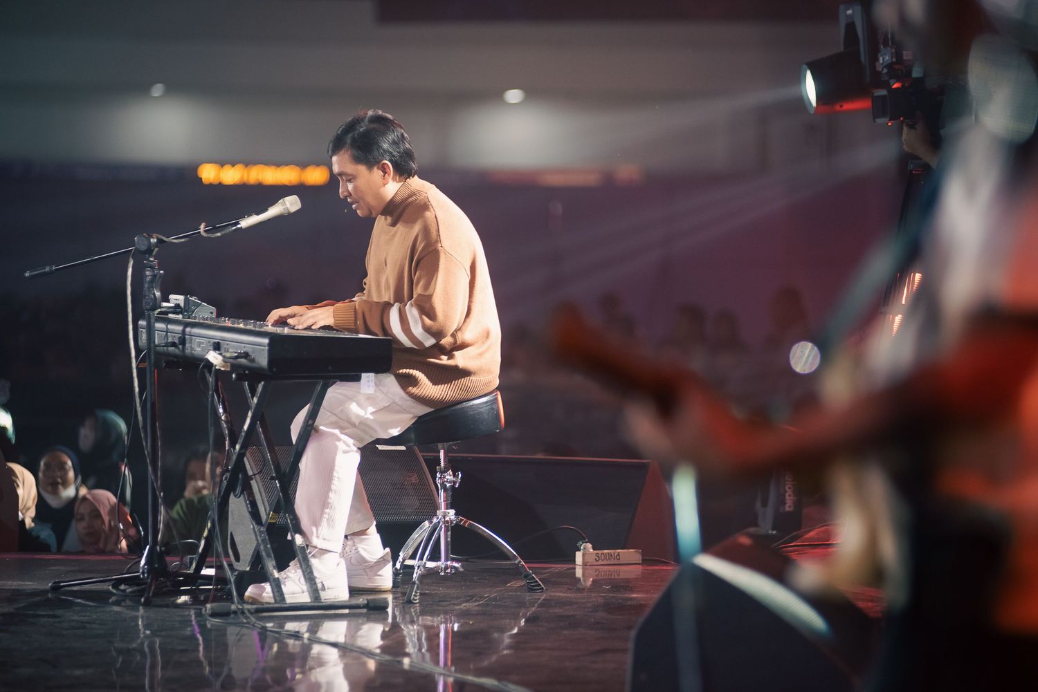 Musician playing keyboard synthesizer on stage with microphone, guitarist visible in foreground at live concert performance.