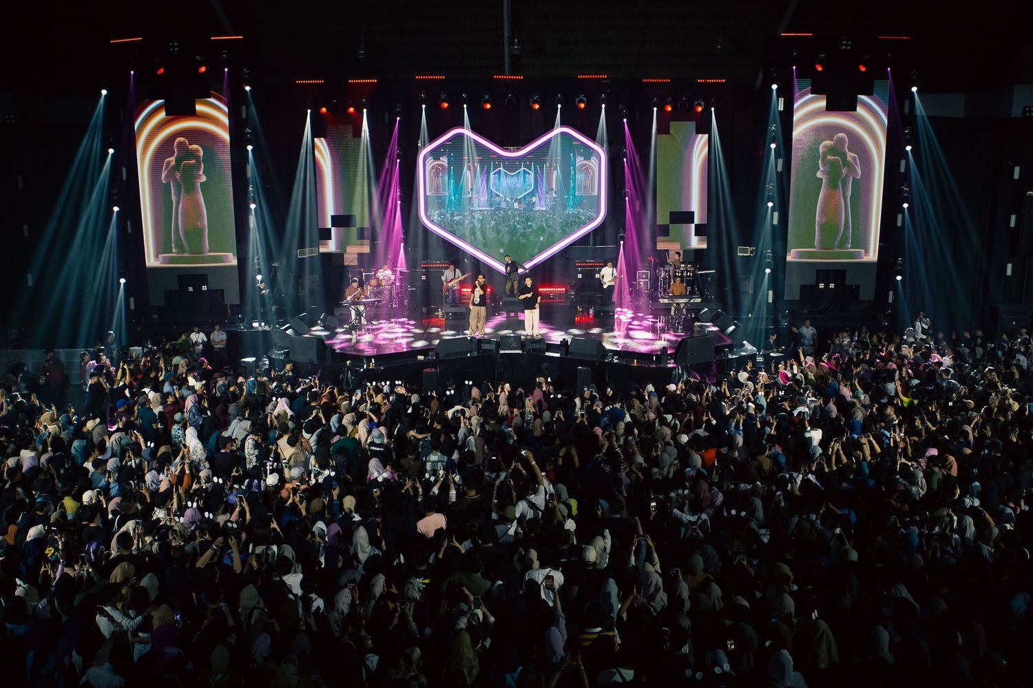 A packed concert crowd cheers as a band performs on a vibrant, pink-lit stage with large decorative screens.