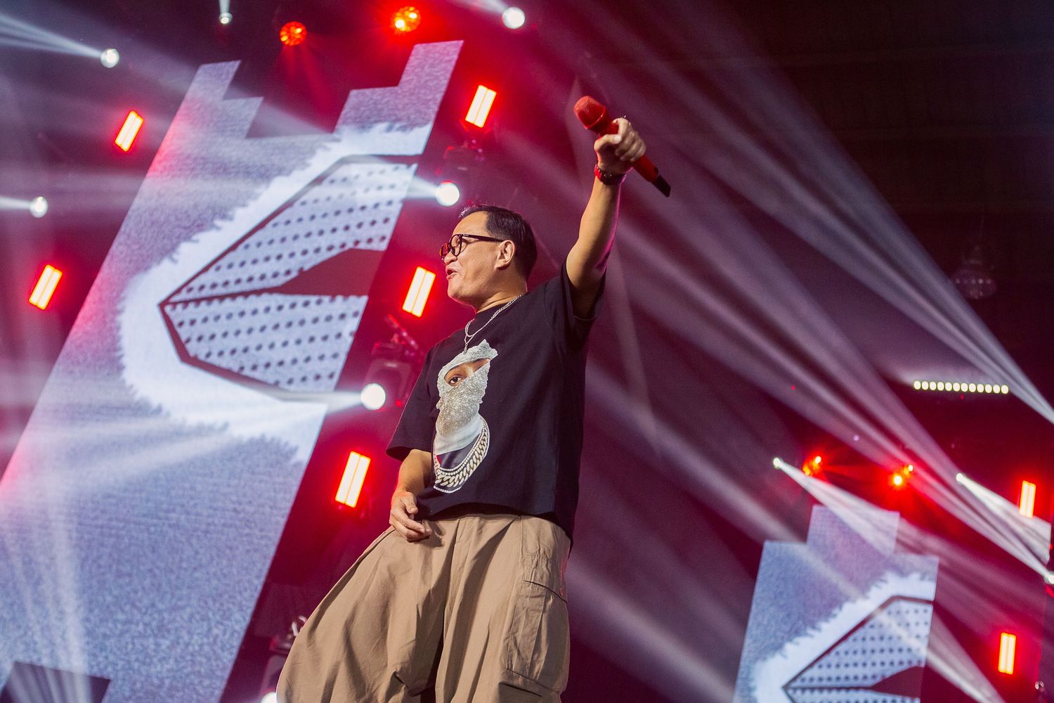 Performer on stage raising microphone under red and white lights at a live concert event.