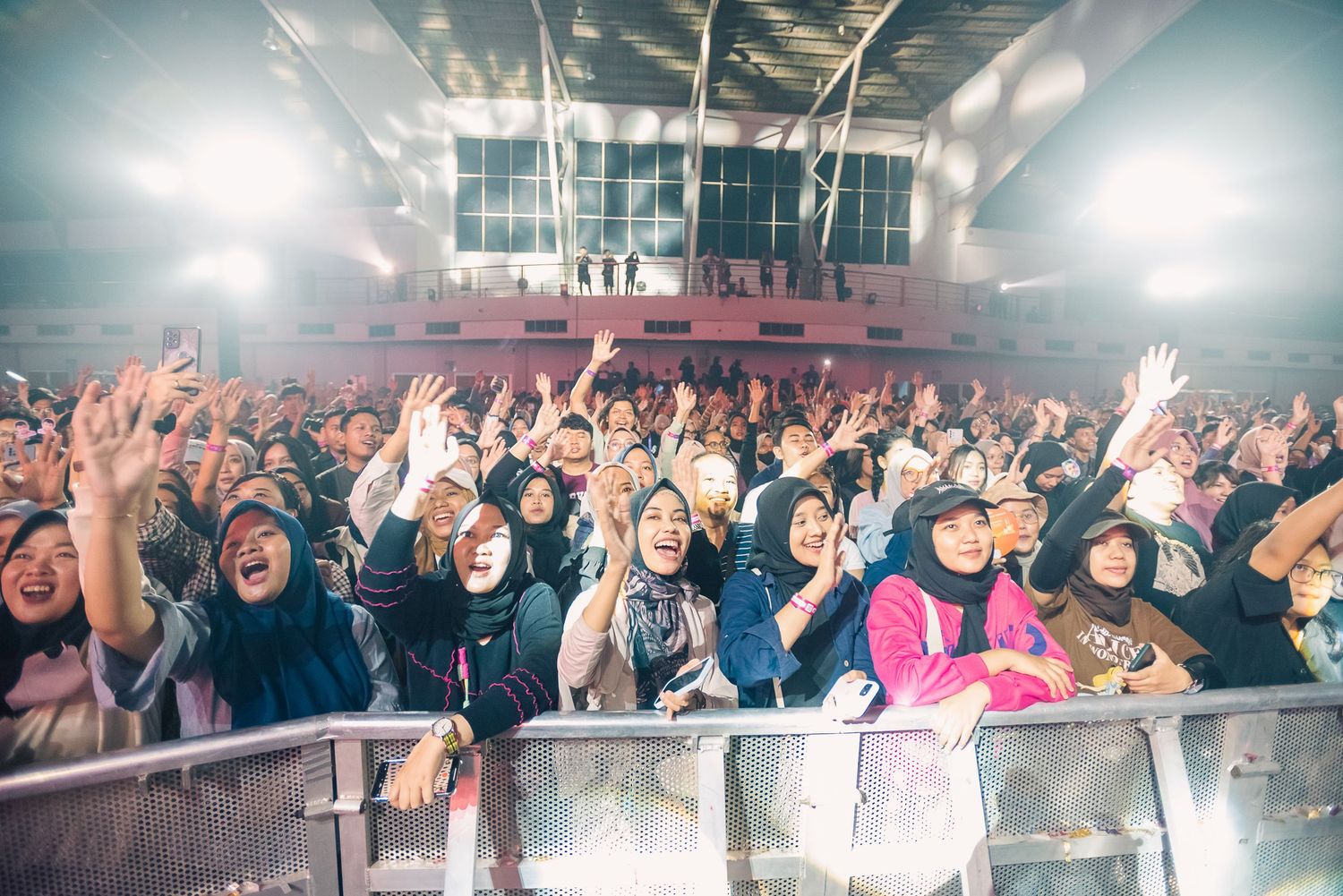 Excited crowd of fans with raised hands at an indoor concert venue, many wearing hijabs, under bright stage lights.