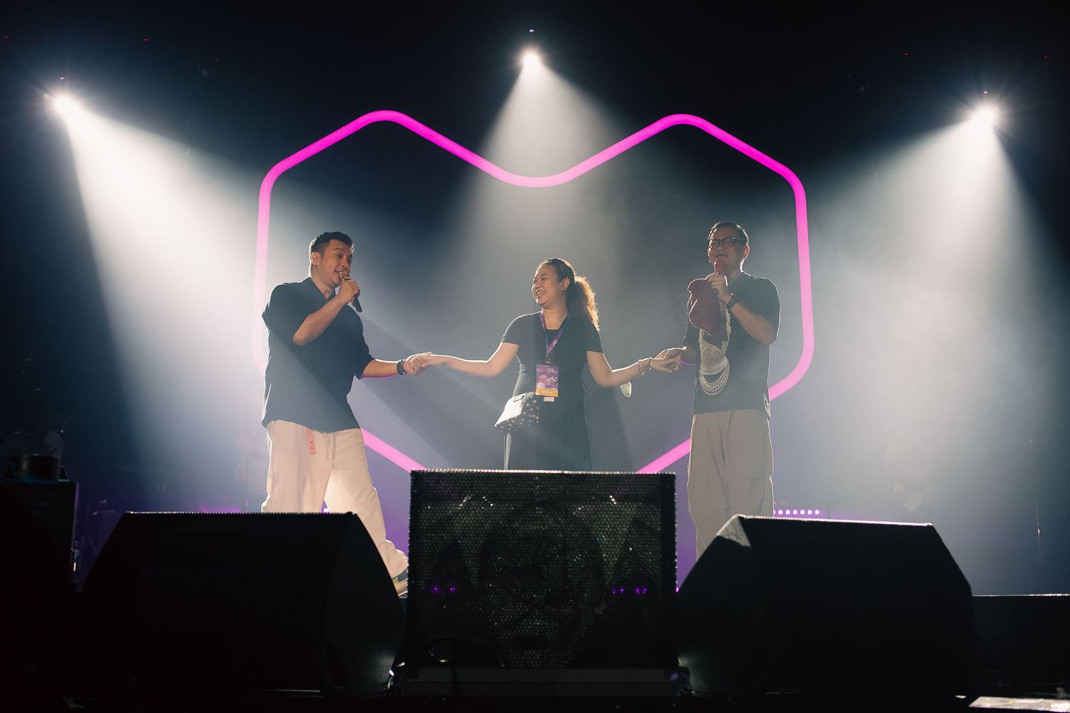 Three performers on stage with pink neon hexagon backdrop and dramatic spotlights at a live music event.