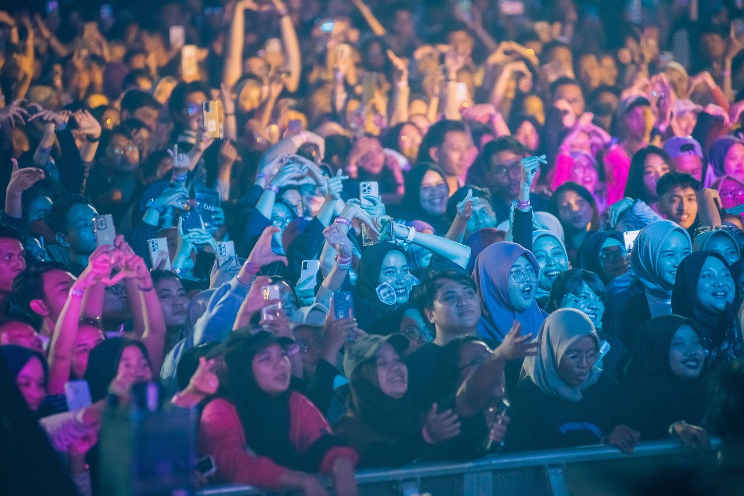 Large concert crowd holding up phones to record a live music event under colorful stage lighting at night.