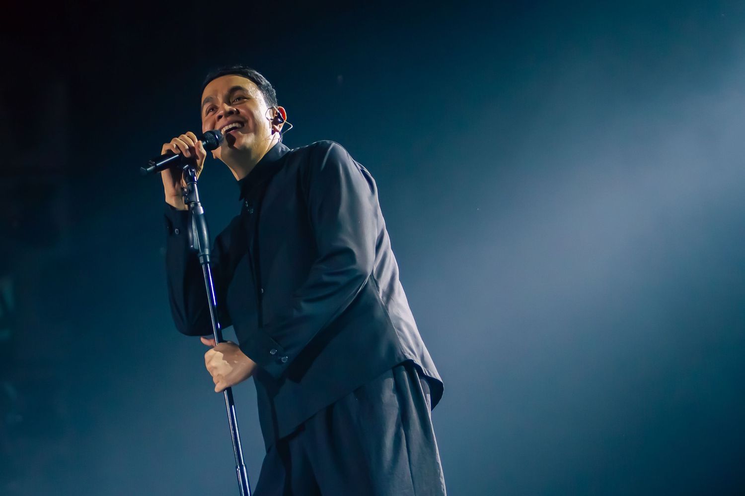 Singer performing on stage holding microphone, wearing dark suit under dramatic blue spotlight in concert venue.