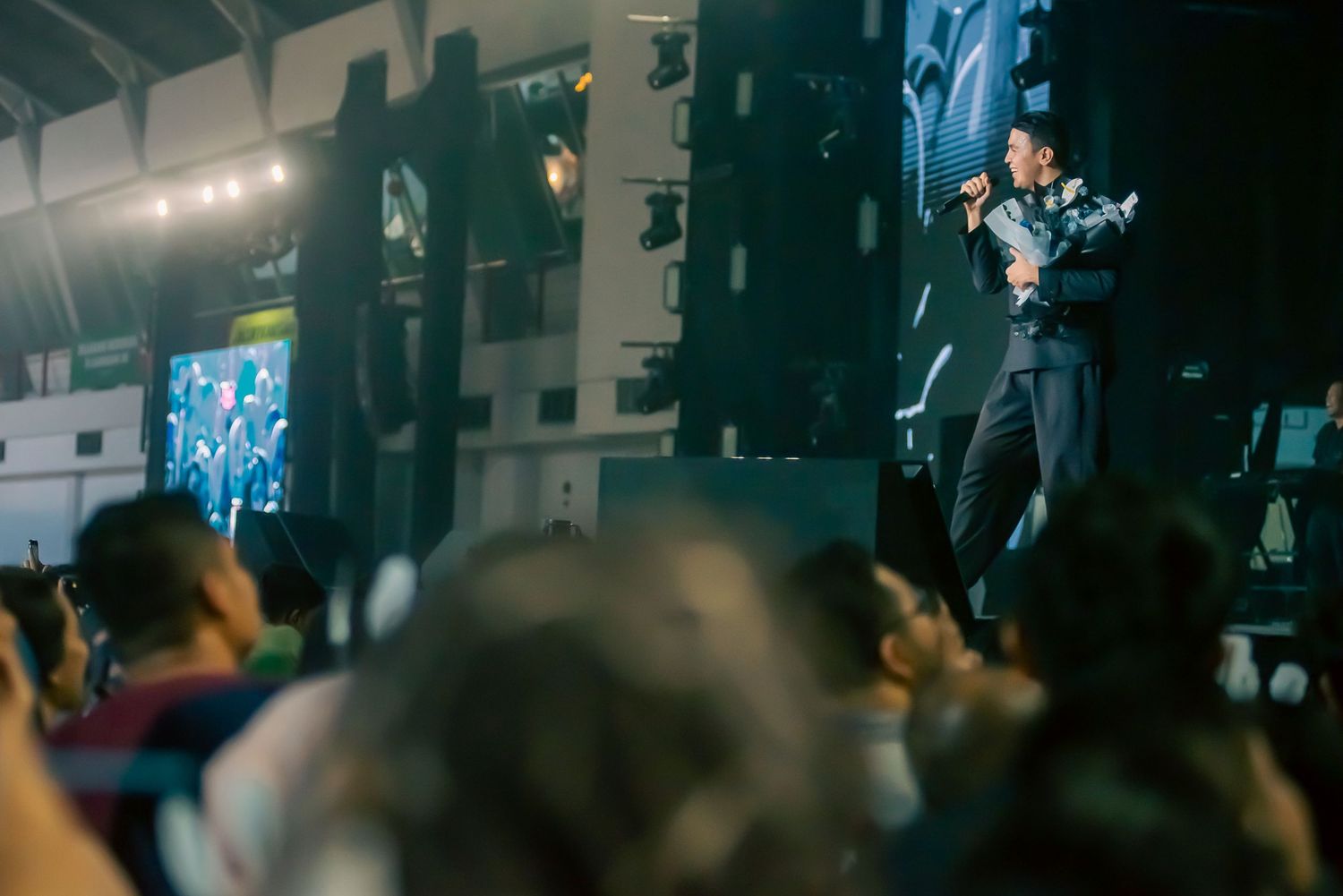 A performer sings on stage with a microphone before a large crowd at an indoor concert event.