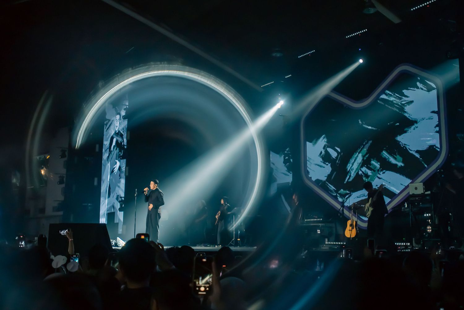 Concert stage with dramatic circular light display and performers silhouetted against glowing screens in dark arena.