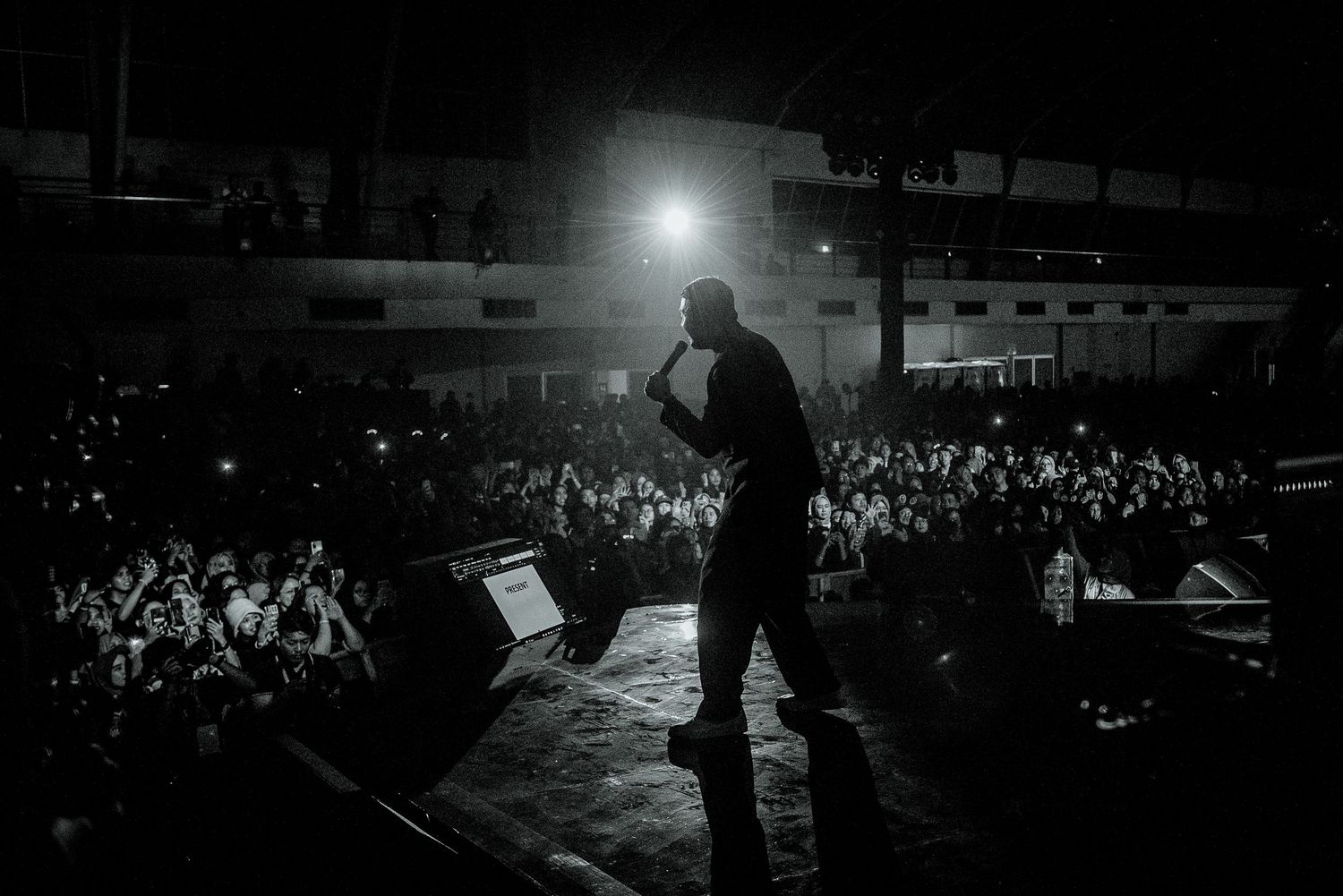Black and white photo of a performer on stage addressing a large crowd at an outdoor concert venue at night.