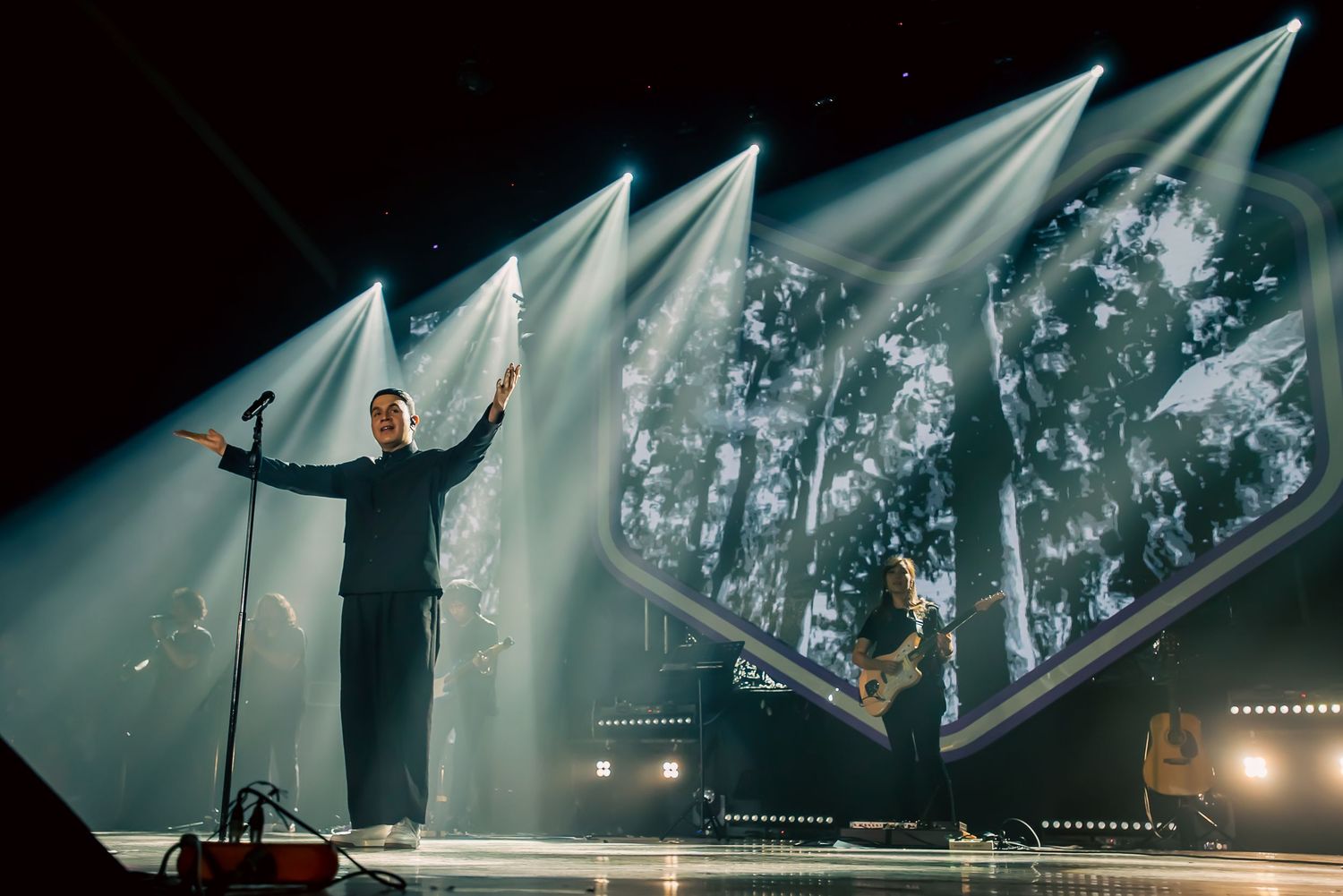 Singer performing on stage with dramatic lighting and large forest projection screen backdrop.