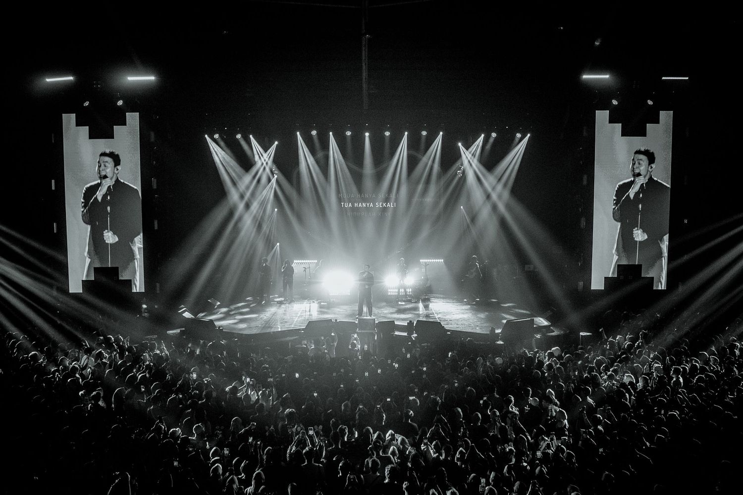 Black and white concert photo showing a performer on stage with dramatic lighting and large video screens.