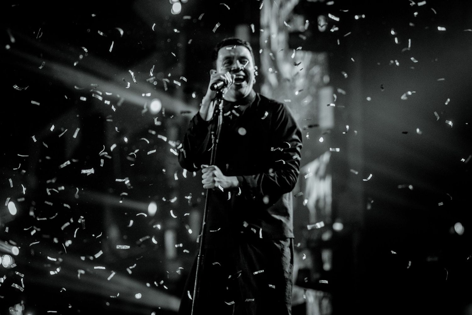 Black and white photo of a male singer performing on stage with confetti falling and dramatic stage lighting.