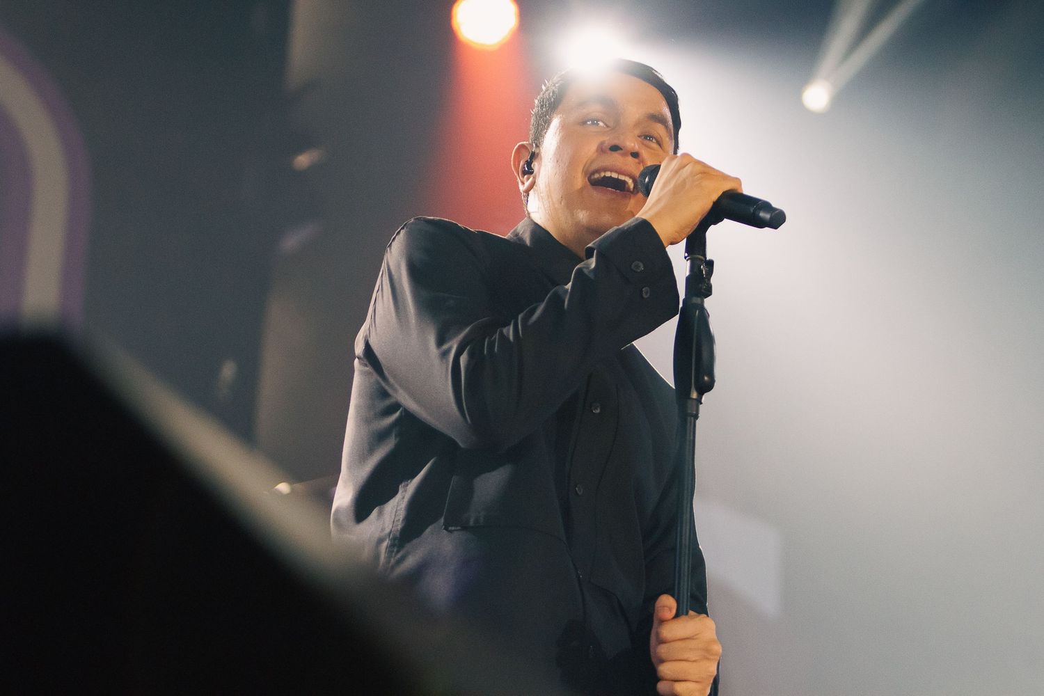 Male singer performing on stage, holding microphone under bright spotlight in dark concert venue.
