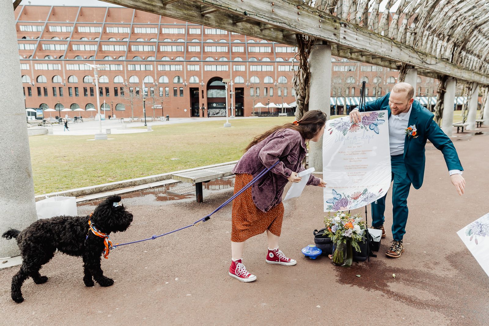 Couple with black dog holding flowers and a sign outdoors near a brick building on a cloudy day.