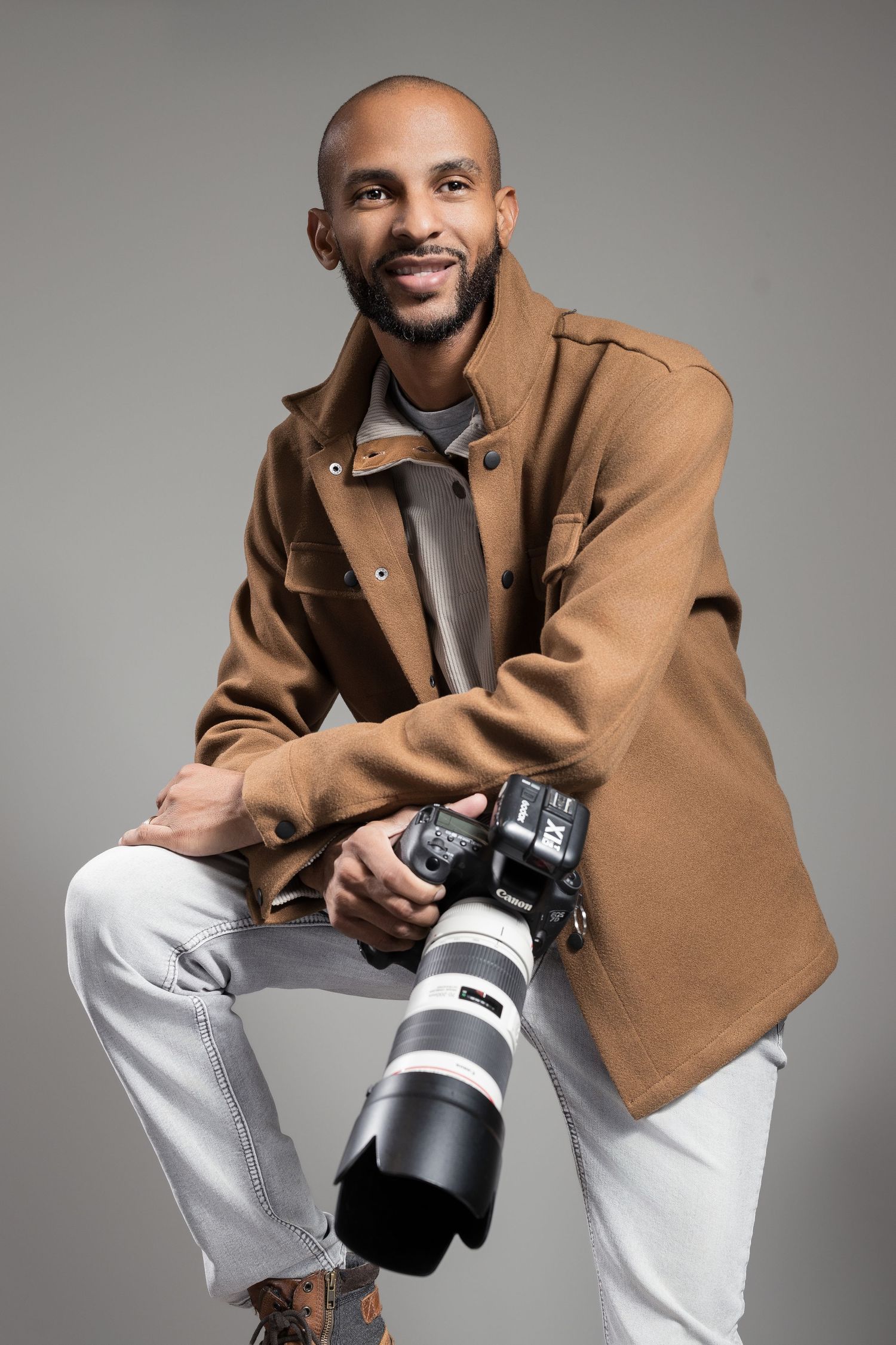 Professional photographer in tan coat holding camera with telephoto lens against grey background.