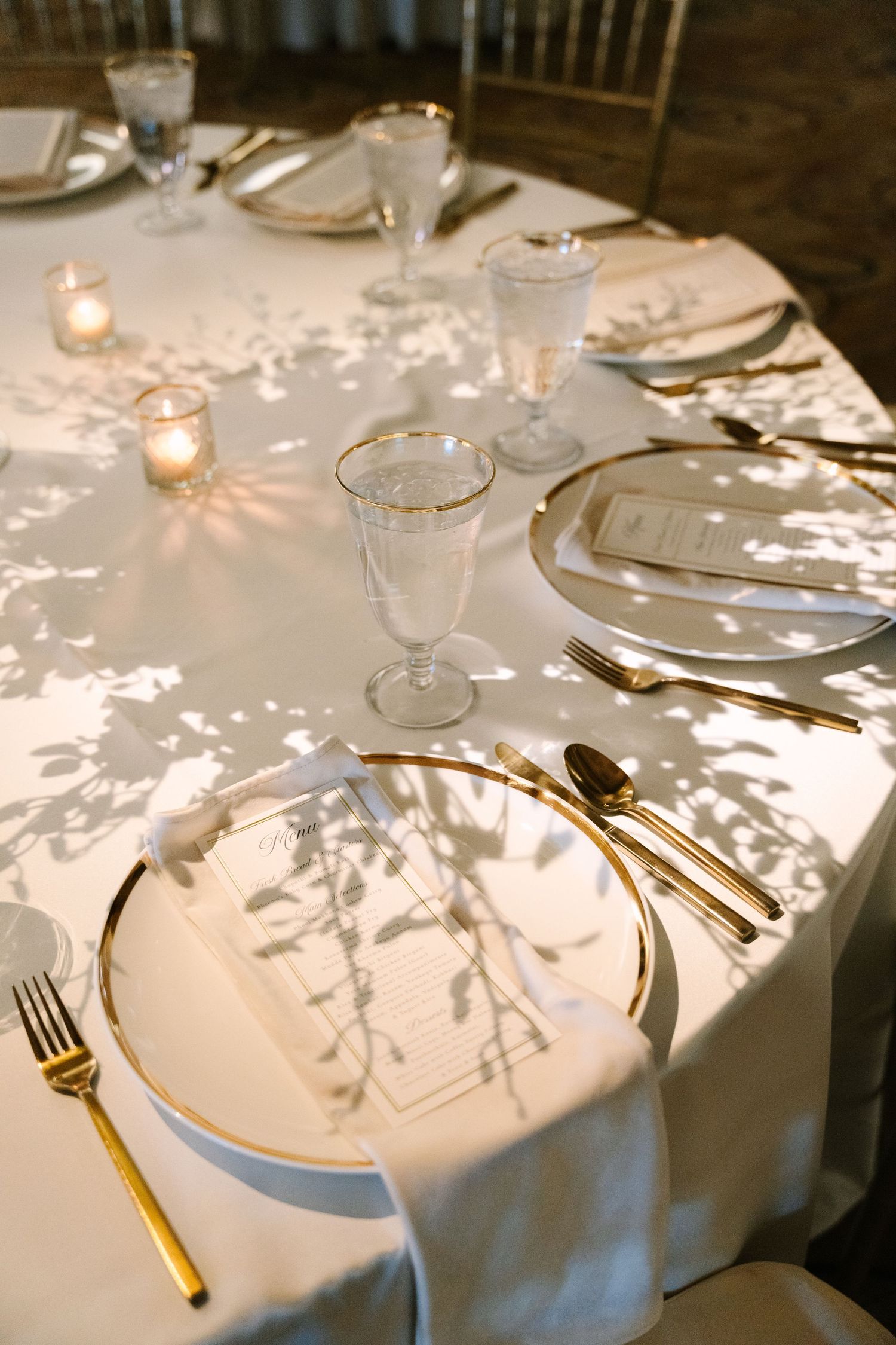 Elegant wedding reception table with gold cutlery, crystal glasses, white linens, and flickering candles casting leafy shadows.