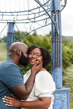 A romantic couple shares a tender moment together under a decorative blue metal gazebo structure outdoors.