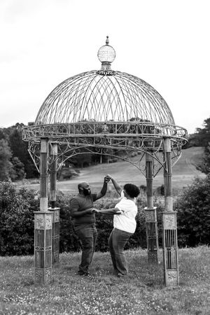 A couple dances together beneath an ornate metal gazebo structure in a field during a romantic photoshoot.