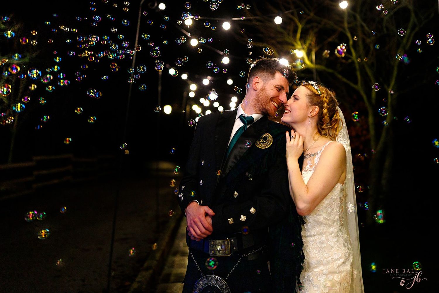 A bride and groom share a romantic moment surrounded by floating bubbles at night, with the groom in a kilt.