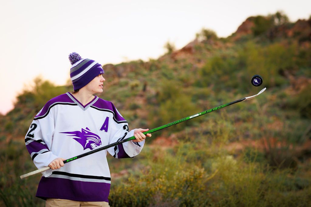 Hockey player in purple jersey and beanie tosses puck with stick outdoors against desert hillside backdrop.