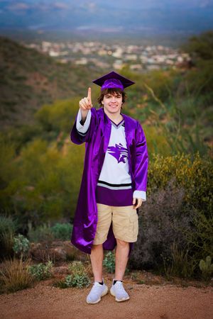 Graduate in purple cap and gown pointing up on desert hillside with city view.