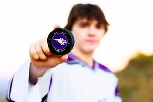 Teen boy holding a purple hockey puck with a bird logo close to the camera outdoors.