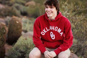Young man smiling outdoors wearing a red Oklahoma Sooners hoodie, seated among desert cacti.