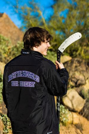 Teen boy in Battlefield Ice Hockey jacket holding a hockey stick outdoors among rocks and trees.