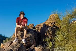 Young man in red shirt and shorts sitting on rocks outdoors with blue sky and green shrubs behind him.