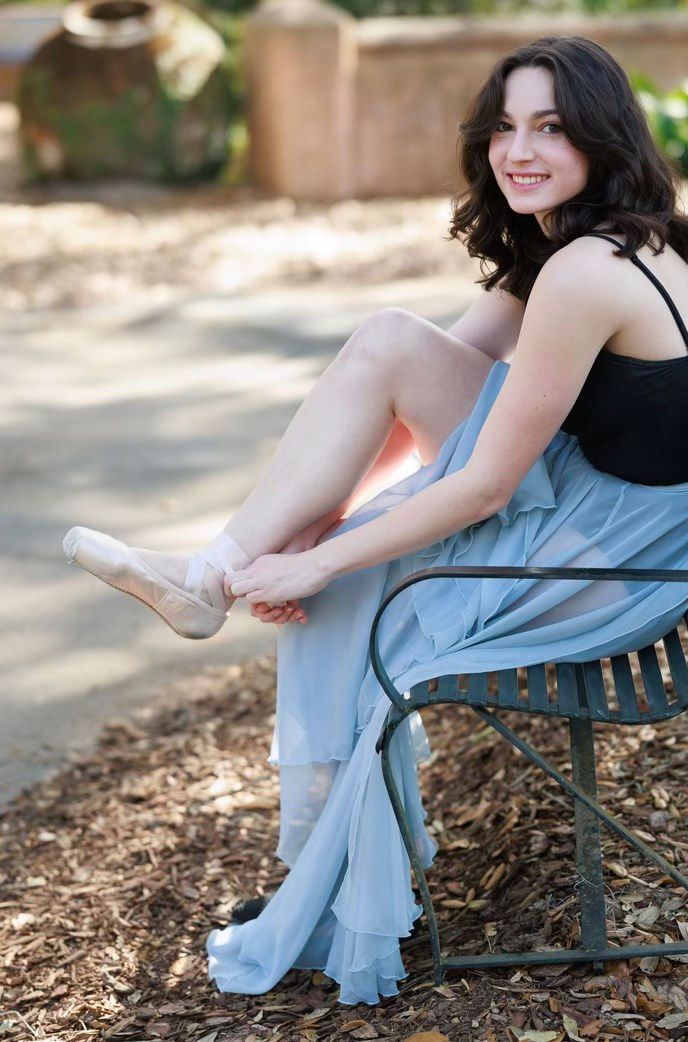 Smiling brunette ballet dancer in blue skirt putting on pointe shoes while sitting on a bench outdoors.