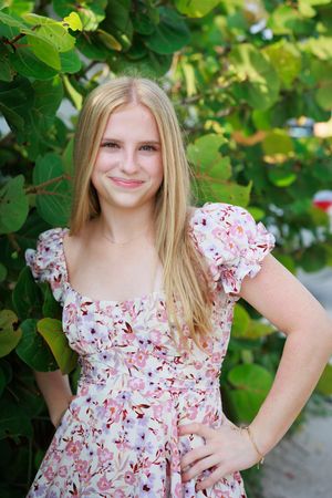 Young blonde girl in a floral pink dress smiling outdoors near lush green tropical foliage.