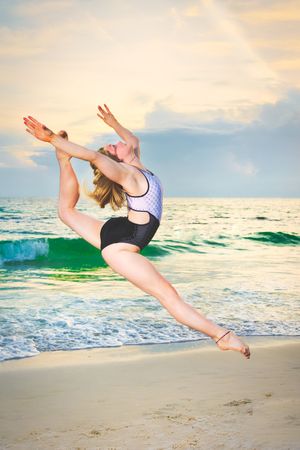 Young female gymnast performing a leap split jump on beach at sunset.