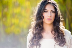 Young woman with long wavy brunette hair and natural makeup poses outdoors with soft green bokeh background.