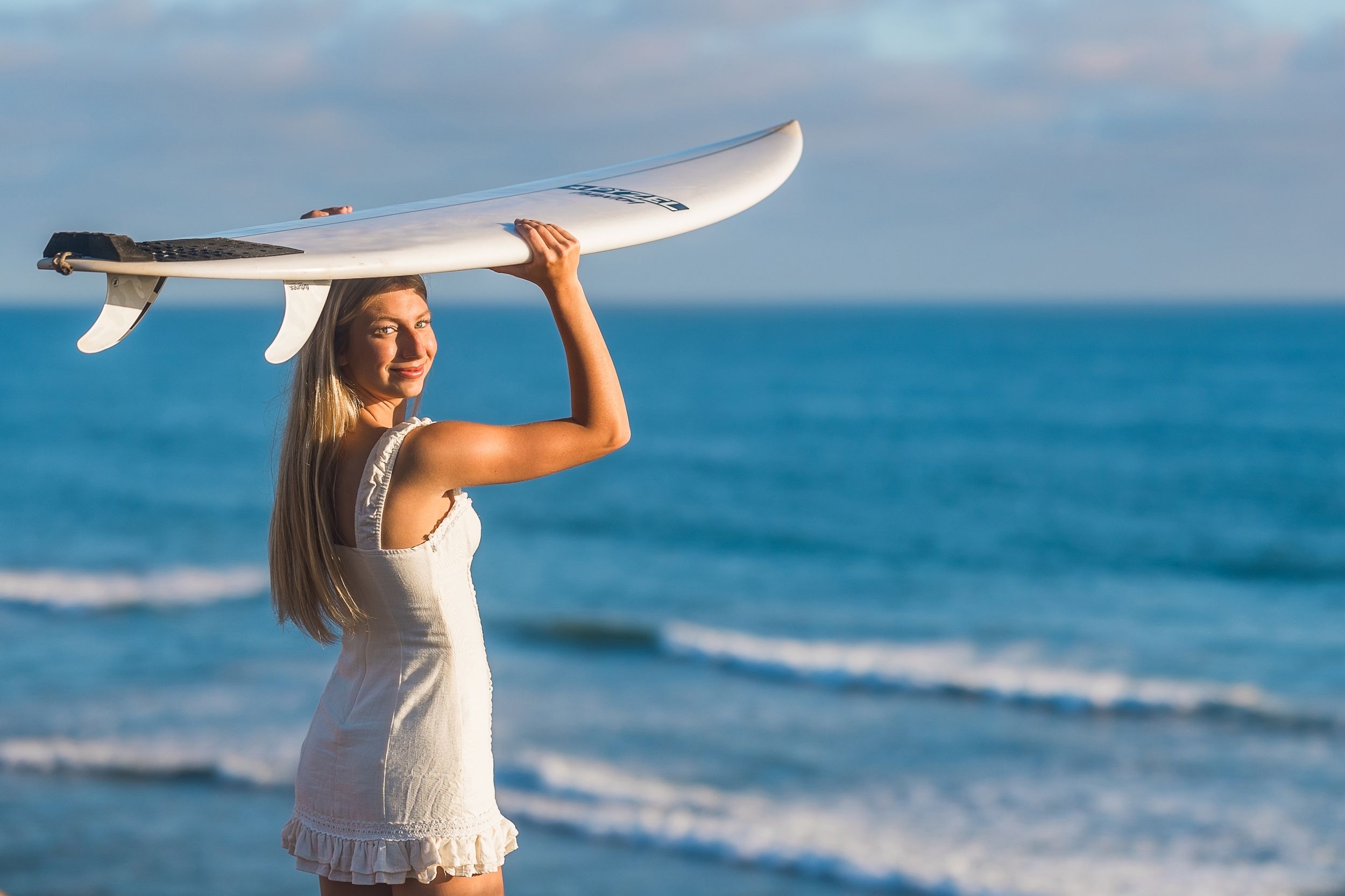 Smiling blonde woman in white dress holding a surfboard overhead on a sunny beach with blue ocean waves.