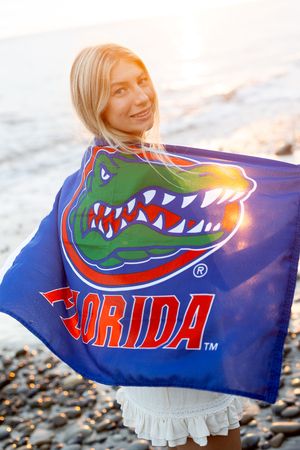 Smiling blonde woman holding University of Florida Gators flag on rocky beach at sunset.