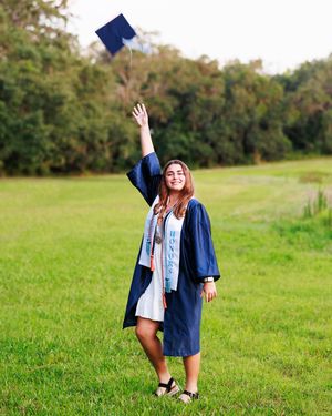 A smiling graduate in navy cap and gown tosses her mortarboard in the air on a lush green field.