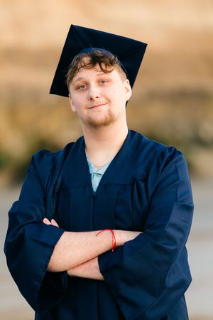 Young male graduate in navy cap and gown stands with arms crossed, smiling confidently outdoors.