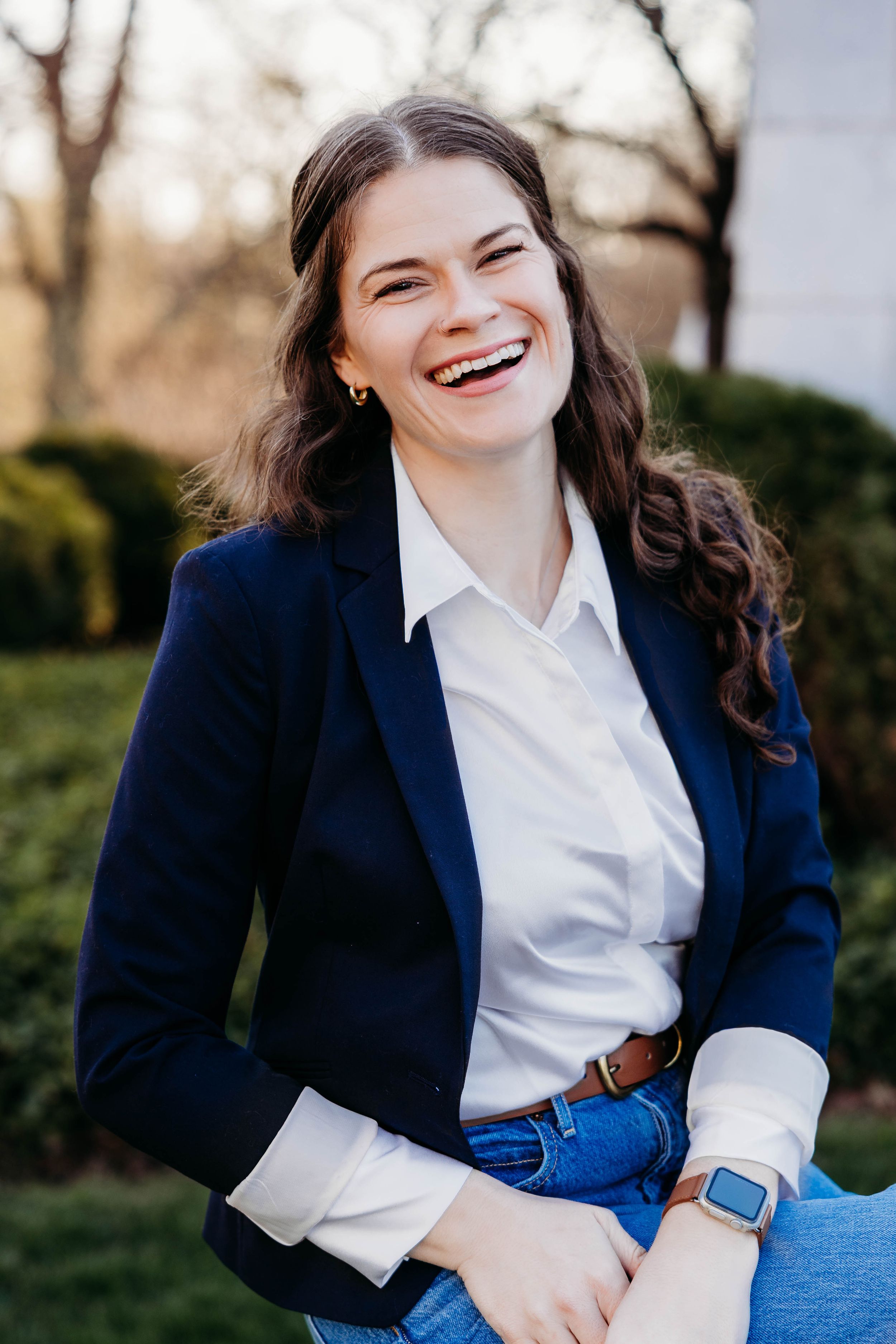 Young woman in navy blazer and white shirt laughing outdoors, wearing a smartwatch, with greenery in background.