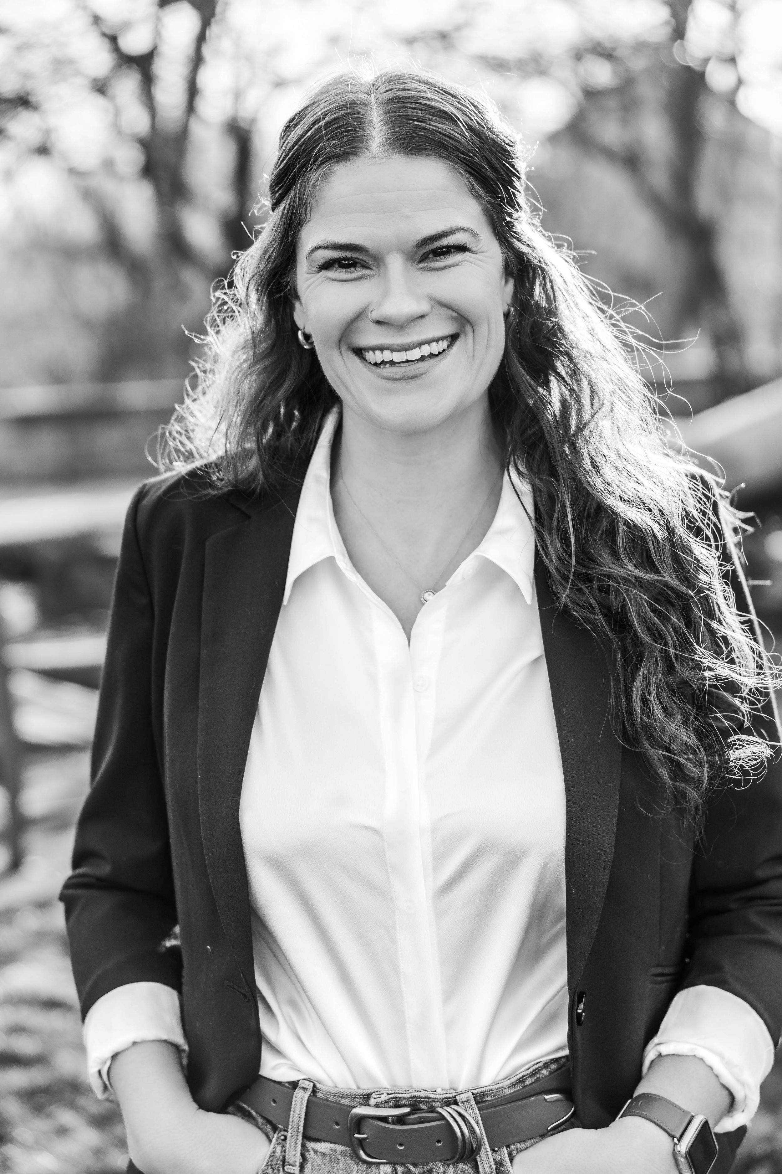 Smiling young woman with long wavy hair wearing a white shirt and black blazer outdoors in a black and white portrait.