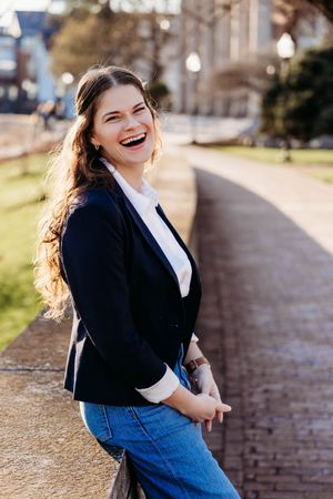 Young woman in navy blazer and jeans laughing outdoors on a sunny day along a brick pathway.