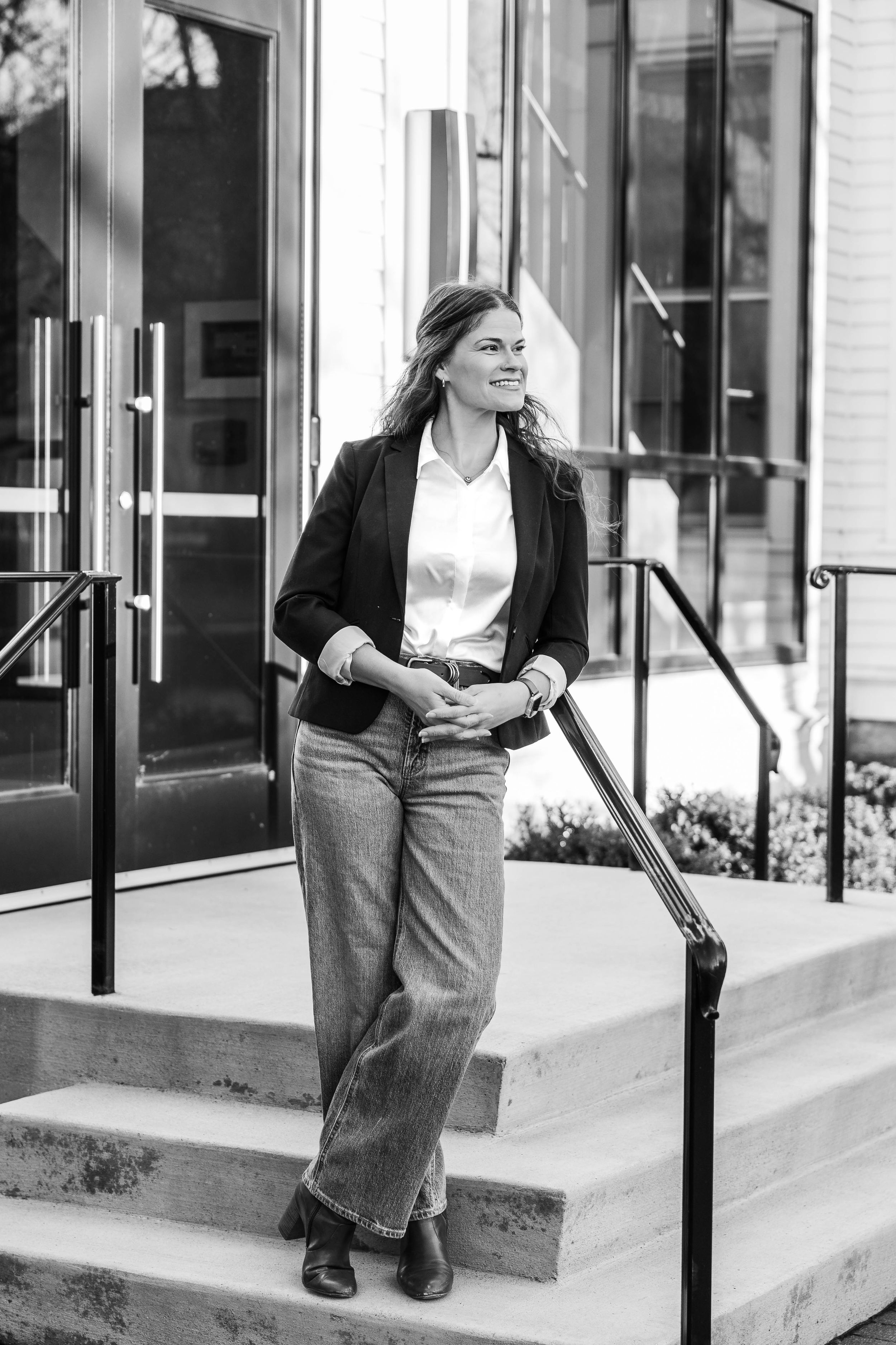 A professional woman in a blazer and jeans smiles on outdoor steps in a black and white portrait photo.