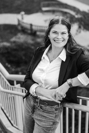 A professional woman in a blazer and jeans smiles on outdoor steps in a black and white portrait photo
