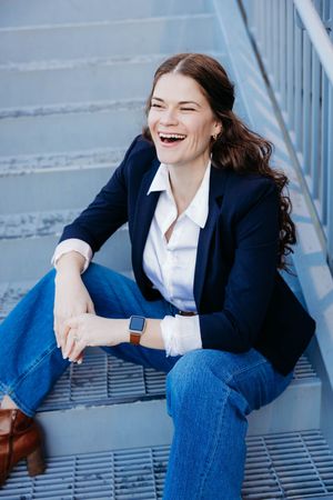 A laughing woman in a navy blazer and jeans sits on outdoor metal steps wearing a smartwatch.