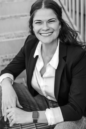 Smiling woman in black blazer and white shirt poses outdoors in a professional black and white portrait photo.