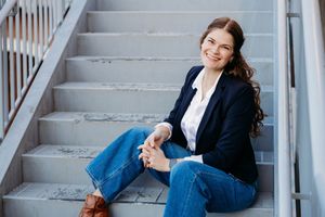 Smiling young woman in navy blazer and jeans sitting relaxed on gray outdoor steps.